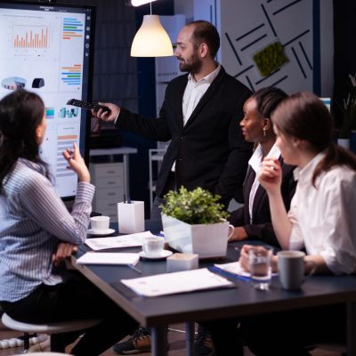 Entrepreneur man brainstorming management strategy working hard in meeting office room late at night. Diverse multi-ethnic business team looking at financial company presentation on monitor.